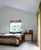 Französisches Bett mit folkloristischer Tagesdecke vor Fenster, seitlich Holztisch mit Leuchte in minimalistischem Dachzimmer