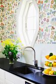 Modern kitchen counter with black worksurface and integrated sink below window in wall with colourful floral wallpaper
