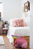 Rosa Plastiktasche und Bastelutensilien in Holzkiste vor schlichtem Bett mit weisser Tagesdecke und Kissen in ländlichem Zimmer