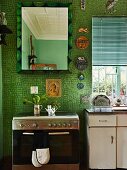 Modern stainless steel cooker and mirror on wall with green mosaic tiles; vintage sink unit with base cabinet to one side below window