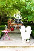 Verschiedene Outdoor Möbel auf podestartigen Stufen im Garten - verspiegelte Kugel auf Boden vor weißem Kunststoff Sessel und Tisch mit Obstschale, dahinter Rattan Sessel mit passendem Schemel