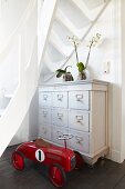 Red, vintage bobby car in front of white-painted chest of drawers under white, winding wooden staircase