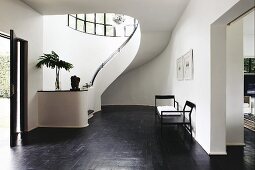 Vorraum mit geschwungener Treppe an gebogener Wand und minimalistische Sitzbank auf dunklem Holzboden, im Vordergrund teilweise sichtbarer Durchgang
