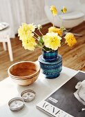 Yellow dahlias in retro ceramic vase in shades of blue and various metal and wood containers on wooden table