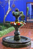 Pool with ornamental, tiered fountain decorated with flowers in courtyard with terracotta tiles and blue wall in background