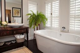 Free-standing bathtub below window, antique washstand with countertop basin below framed mirror and fern on plant stand in corner