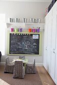 Children's table and chairs with striped loose covers below drawings on wall-mounted blackboard; colourful metal buckets hanging from bracket shelves above