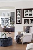 Orchidee und Tablett auf grau bezogenem Pouf gegenüber hellem Sessel, seitlich an Wand gerahmte Fotos