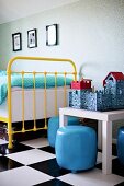 Yellow, vintage, metal-framed bed and pale blue pouffes around toy castle on small table on chequered floor