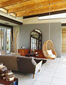Designersofa, Hängekorbstuhl und ein Standspiegel mit Fenstersprossen in gefliestem, offenem Wohnraum