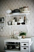 Crockery on bracket shelves on patterned wallpaper above white-painted half-height cabinet