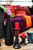 Arrangement of various wooden candlesticks and black ceramic vase on wooden terrace in front of comfortable couch