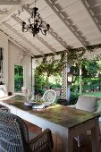 Chandelier, wicker chairs and battered table on roofed, country-house terrace; view of garden through climber-covered white wooden trellising