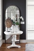 Orchids on white, round side table in front of arched, full-length mirror leaning against dark wall