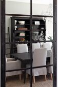View through glass door with black lattice frame of dark wood dining table, chairs with pale grey loose covers and antique wooden cabinet