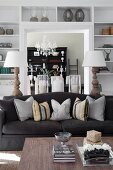 Coffee table, black sofa with arranged scatter cushions, two table lamps and white fitted shelves surrounding doorway in background