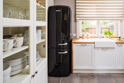 Schwarzer Retro Kühlschrank in Landhausküche, Küchenzeile mit Spülbecken vor Fenster und halbgeschlossenes Rollo mit weissbraunen Streifen, vorne Vitrinenschrank