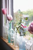 Verschiedene Glasgefässe mit zart pinkfarbenen Tulpen und Pfingstrosen auf schmalem Fensterbrett