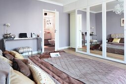 Elegantes Schlafzimmer mit Satindecken und -kissen auf Doppelbett und Kleiderschrank mit Spiegeltüren; im Hintergrund offene Tür zum Bad Ensuite