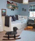 Schaukelschaf neben kariertem Teppich in weiss-blauen Pastellfarben, vor weißem Einzelbett mit hoher Seiten- und Rückenlehne, im Kinderzimmer
