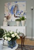 Blue glass vase of roses on coffee table, houseplants and bust on sideboard below modern artwork in background