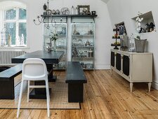 Esstisch und Sitzbänke aus schwarz gestrichenem Holz und Kinderstuhl; Sideboard im Antikstil und moderne Glasvitrinen im Hintergrund