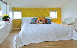 Double bed with headboard, white bed linen and colourful scatter cushions against accent wall with yellow, bird-patterned wallpaper; white designer chair to one side