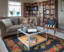 Coffee table on floral rug, sofa and antique chair in front of glass-fronted, L-shaped bookcase