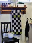 Chequered towel on towel rail with ornaments on wooden shelf above black-painted wooden chair
