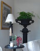 Table lamp with white lampshade on round side table, and foliage plants in black, antique urn on plinth in background