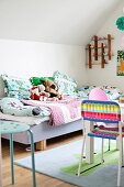 Child's bed with colourful blankets, pillows and soft toys; colourful plastic chair at play table in foreground