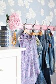 Denim clothing hung from hooks on floral wallpaper; hydrangea bloom in retro vase on white chest of drawers