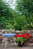 White, delicate metal chair with blue seat cushion next to red geraniums on stone floor in front of low garden wall with view into garden