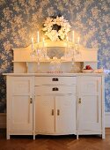Crystal candelabra with lit candles on white-painted cabinet with top section against wallpaper with white floral pattern on blue background