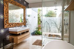 Teilweise sichtbare Badewanne vor verglastem Duschbereich und Blick auf Terrasse, seitlich Waschtisch unter Spiegel mit holzgeschnitztem Rahmen an tapezierter Wand