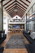 View from lounge area with black leather sofa set and vintage rug to dining area and mezzanine below exposed roof structure in contemporary home