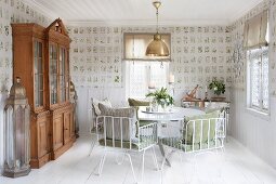 weiße Metallstühle mit Kissen um rundem Tisch und Vitrinenschrank in Esszimmer mit botanischem Tapetenmuster und weißem Dielenboden