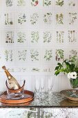 Sparkling wine in wine cooler and glasses on console table against botanical wallpaper