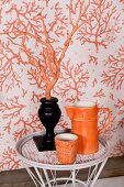 Orange jug and beaker next to orange branch in black vase in front of patterned wallpaper