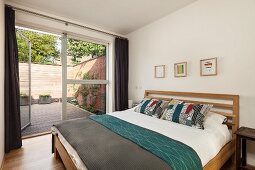Double bed with wooden frame in simple bedroom with open terrace door in glass wall