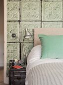 Tischleuchte auf Nachtkästchen aus Metall neben Bett mit türkisfarbenem Kissen, an Wand alte 3D-Fliesen