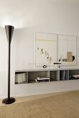 Modern standard lamp in black metal next to white floating shelf on wall below artworks in minimalist interior