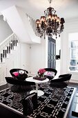 Classic armchairs made from metal slats around white Tulip Table on black and white patterned rug under chandelier next to staircase