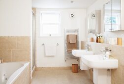 Pale bathroom with twin sinks on projecting wall below mirrored cabinets and sand-coloured tiles on floor and walls