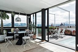 Seating area with delicate, white metal chairs and arc lamp in conservatory with open sliding door leading to roof terrace with view of city