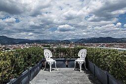 Two white, vintage, Rococo-style metal chairs positioned in corners of planted roof terrace with magnificent panoramic view