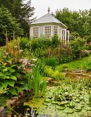 Blick über Teich mit blühenden Wasserpflanzen, auf Gartenpavillon aus Holz mit Sprossenfenstern in idyllischem Garten