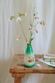 Grüne Vintage Vase mit Wiesenblumen (Taubenkropf-Leimkraut) auf rustikalem Holzhocker, vor weißem Leinenvorhang