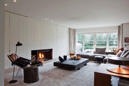 Modern interior in Scandinavian style - comfortable lounge with open fire, low coffee table made from dark wood, corner sofa and reading chair below black standard lamp in foreground