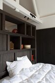 Bed with white bed linen against dark wooden partition shelving below gabled ceiling